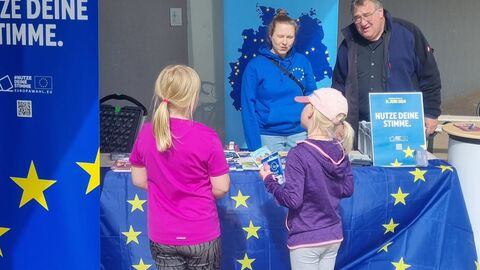 Zwei Mädchen informieren sich zur Europawahl. Zwei Mädchen stehen vor dem Infostand zur Europawahl, am Stand stehen Regierungspräsident Mark Weinmeister und eine Mitarbeiterin des Europe Direct NordOstHessen.
