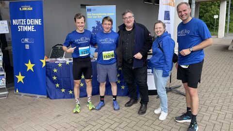 Regierungspräsident Mark Weinmeister mit dem Team des Europe Direct NordOstHessen Regierungspräsident Mark Weinmeister steht mit vier Mitarbeiterinnen und Mitarbeitern, die teilweise Laufkleidung tragen, vor dem Stand des Europe Direct NordostHessen.