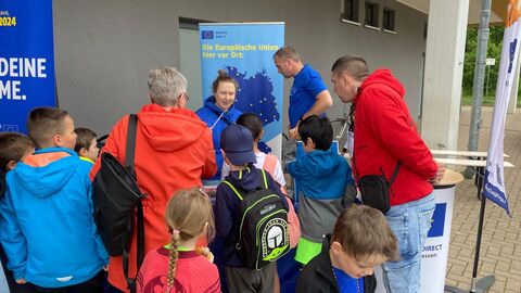 Reges Interesse am Informationsstand zur Europawahl Zwei Erwachsene und viele Kinder stehen vor dem Stand des Europe Direct NordOstHessen, der von einer Mitarbeiterin und einem Mitarbeiter betreut wird.der