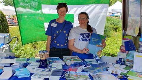 Zwei Mitarbeiterinnen der Staatskanzlei bzw. des Europe Direct Rhein-Main am Infostand zur Europawahl Zwei Frauen stehen hinter einem mit Infomaterial belegten Stand mit einer EU-Flagge und blicken in die Kamera.