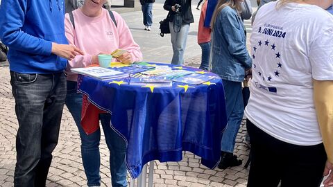 Infomaterial zur Europawahl Ein Mann und eine Frau stehen an einem Stehtisch und betrachten sich Infomaterial zur Europawahl. Im Hintergrund sind weitere Menschen zu sehen.
