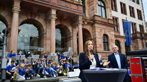 Rede von Landtagspräsidentin Astrid Wallmann Landtagspräsidentin Astrid Wallman hält eine Rede an einem Stehtisch auf der Bühne. Hinter ihr steht Europaminister Manfred Pentz. Im Hintergrund sind Mitglieder der Band der Diltheyschule zu sehen, die auf den Stufen des Rathauses sitzen.