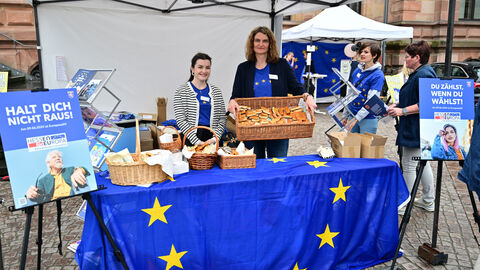 Der Stand der Hessischen Staatskanzlei Zwei Mitarbeiterinnen der Hessischen Staatskanzlei stehen hinter einem Tisch, der mit einer Europafahne dekoriert ist. Darauf stehen Körbe mit Europabackwaren. Eine der beiden Frauen hält einen weiteren, größeren Korb in Richtung der Kamera. Links und rechts neben dem Tisch stehen Staffeleien, auf denen Wahlpalakte präsentiert werden. Das linke Plakat trägt die Aufschrift „Halt dich nicht raus!“, verbunden mit einem Foto eines älteren Herren und dem hessischen Europawahllogo mit der Aufschrift „Hessen für E