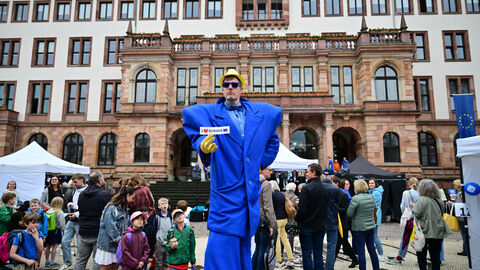 Der europablaue Happy Agent wirbt für Europa Ein Stelzenläufer in einem übergroßen, europablauen Anzug mit breiten Schultern steht auf dem Schlossplatz inmitten verschiedener Festbesuchender. Er trägt eine blaue Brille sowie einen gelben Hut und gelbe Handschuhe. Seine linke Hand hält er hinter dem Rücken, in der rechten Hand hält er einen Aufkleber mit der Aufschrift „I love Europe“ und einer Europaflagge, den er in Richtung der Kamera streckt. Das Wort „love“ wird dabei durch ein rotes Herz ausgedrückt. Links neben ihm steht eine Gruppe Grundschulki