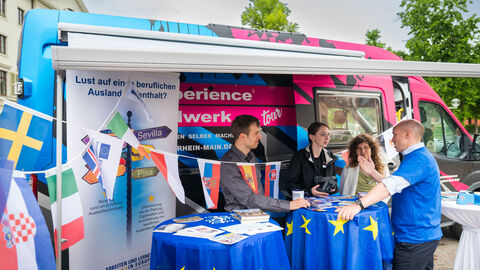 Europaminister Manfred Pentz im Gespräch am Stand der hessischen Handwerkskammern Europaminister Manfred Pentz steht am Stand der hessischen Handwerkskammern an einem Stehtisch, der mit einer Europaflagge dekoriert ist. Darauf liegen verschiedene Informationsmaterialien aus. Ein weiterer Tisch mit Materialien steht links daneben. Herr Pentz trägt ein blaues Europa-T-Shirt mit einem gelben Sternenkreis und spricht mit einem Vertreter und zwei Vertreterinnen der Handwerkskammern. Hinter ihnen steht ein Transporter, der für die Arbeit der Handwerkskammern und berufliche Auslandsaufenthalte