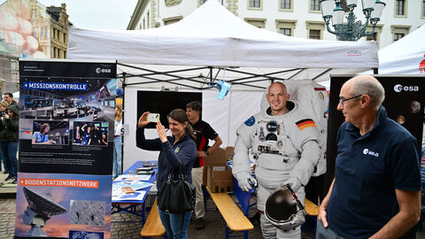 Der Stand der Europäischen Weltraumorganisation ESA Vor dem Stand der Europäischen Weltraumorganisation ESA stehen links und rechts jeweils ein Roll-up-Plakat mit Informationen zur Arbeit der ESA. Vor dem rechten Plakat steht ein Mitarbeiter der ESA, der zu einer Frau blickt, die inmitten des Standes steht und mit ihrem Handy Fotos macht. Neben ihr steht ein lebensgroßer Papp-Aufsteller des deutschen Astronauten Alexander Gerst, mit dem Besuchende Fotos aufnehmen können. Im Hintergrund sind zwei Festzeltgarnituren zu sehen, auf denen Bastel- und Informations