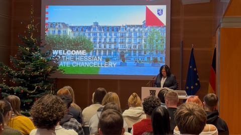 Staatssekretärin Karin Müller steht an einem Rednerpult und begrüßt eine Gruppe Jugendlicher, die in Reihen vor ihr sitzen. Im Hintergrund ist auf einer Leinwand eine Präsentation zu sehen, die ein Foto der Hessischen Staatskanzlei zeigt.